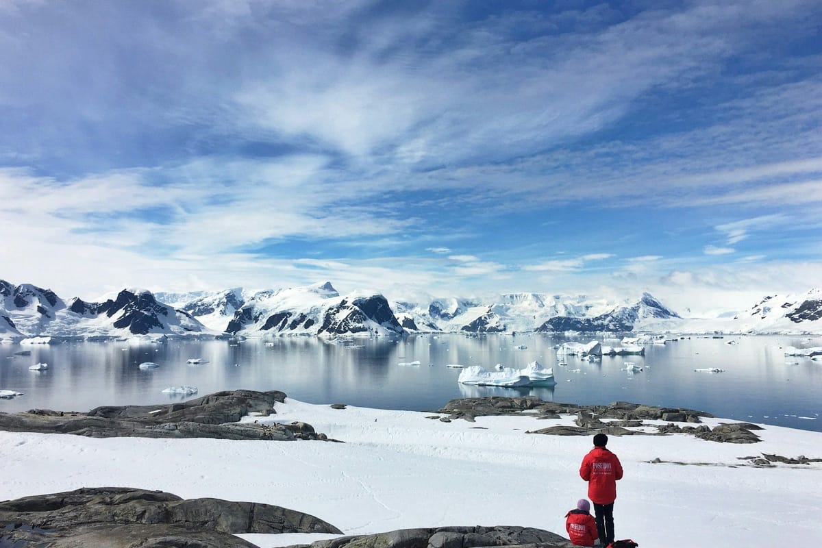 Two people in red parkas overlooking an Antarctic bay filled with icebergs and surrounded by snow-covered mountains.