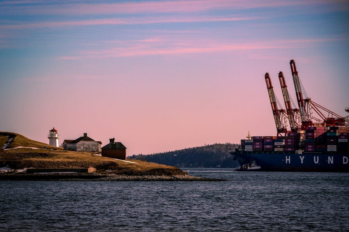 Container ship passing a lighthouse at the entrance to a harbour beneath a pink evening sky.