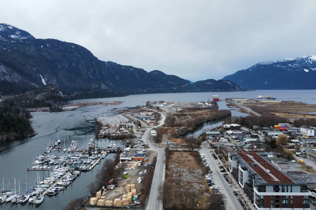 Aerial view of a marina and coastal industrial area surrounded by steep, snow-dusted mountains and a sheltered inlet.