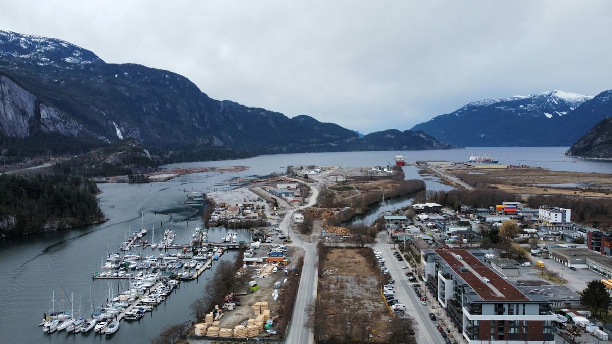 Aerial view of a marina and coastal industrial area surrounded by steep, snow-dusted mountains and a sheltered inlet.