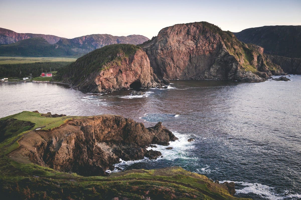Rugged coastal cliffs surrounding a sheltered bay, with a small red house near the shoreline and mountains in the distance.