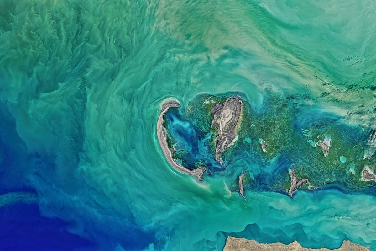 Satellite view of shallow turquoise waters swirling around small islands, sandbanks, and coral reefs.