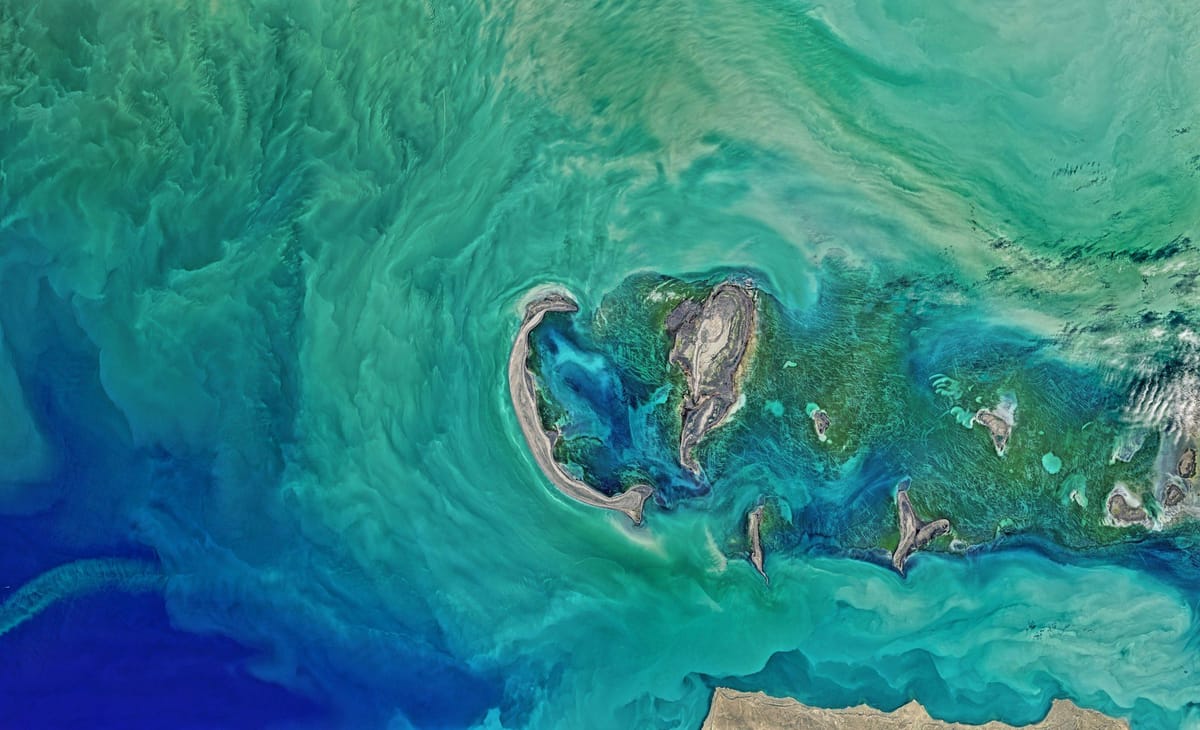Satellite view of shallow turquoise waters swirling around small islands, sandbanks, and coral reefs.