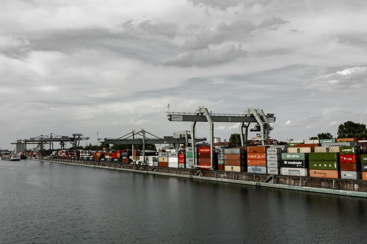 Shipping containers and cargo cranes lining a busy port terminal beside the water.
