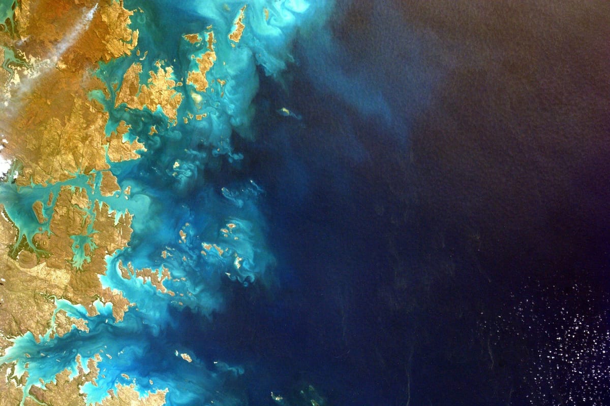 Satellite view of a rugged island coastline, with turquoise shallows fading into deep blue ocean.