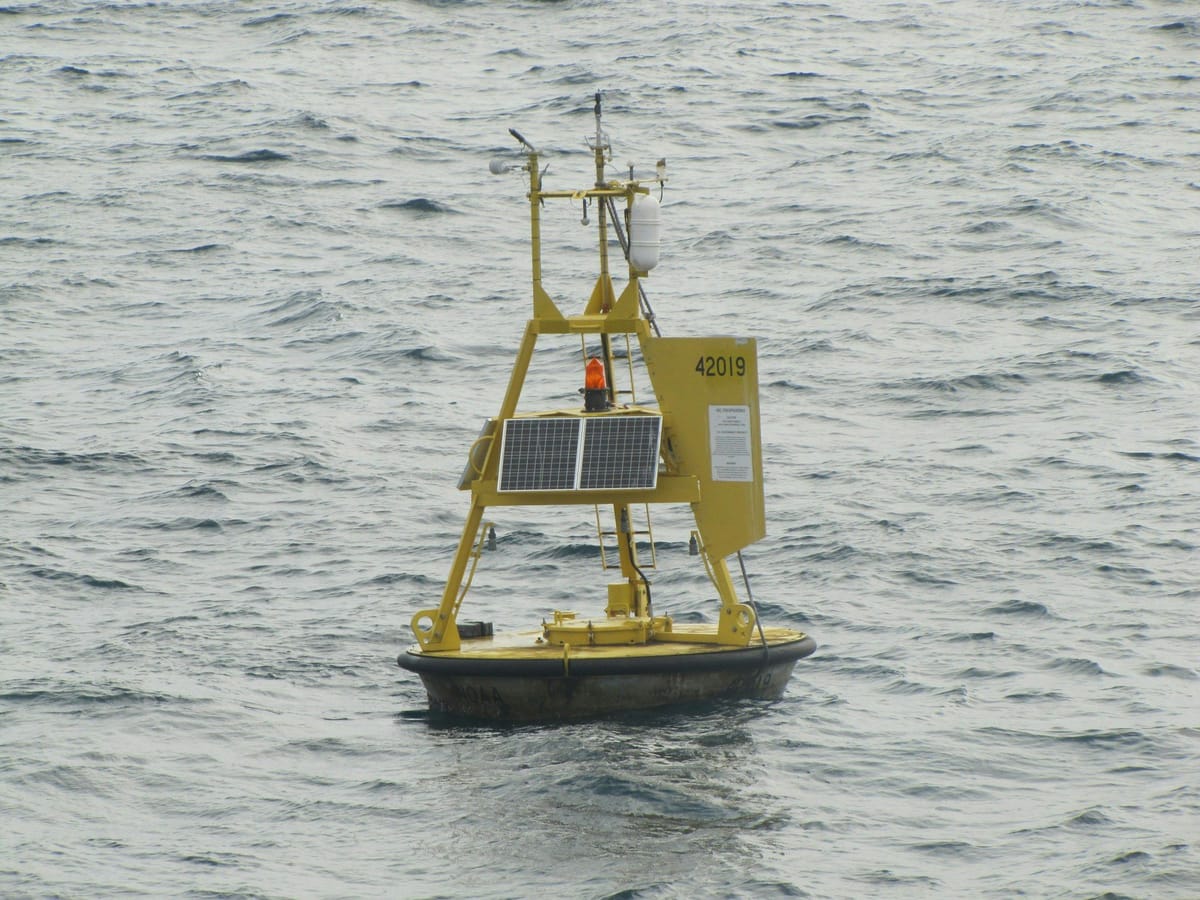 Yellow ocean-monitoring buoy with solar panels and instruments floating in choppy water.