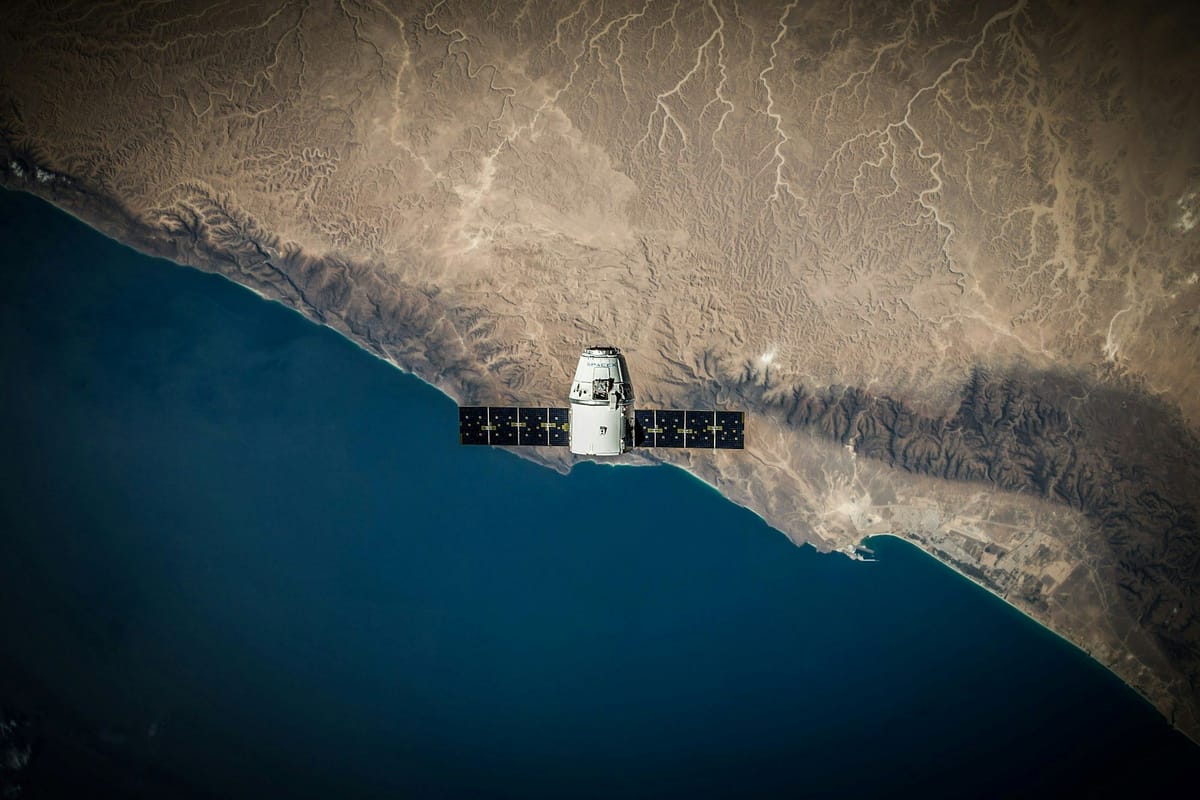 Spacecraft with solar panels orbiting above a rugged desert coastline and deep blue ocean.