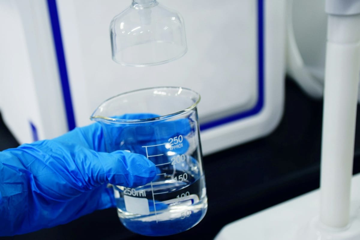 Blue-gloved hand holding a glass beaker of clear liquid beneath laboratory equipment.