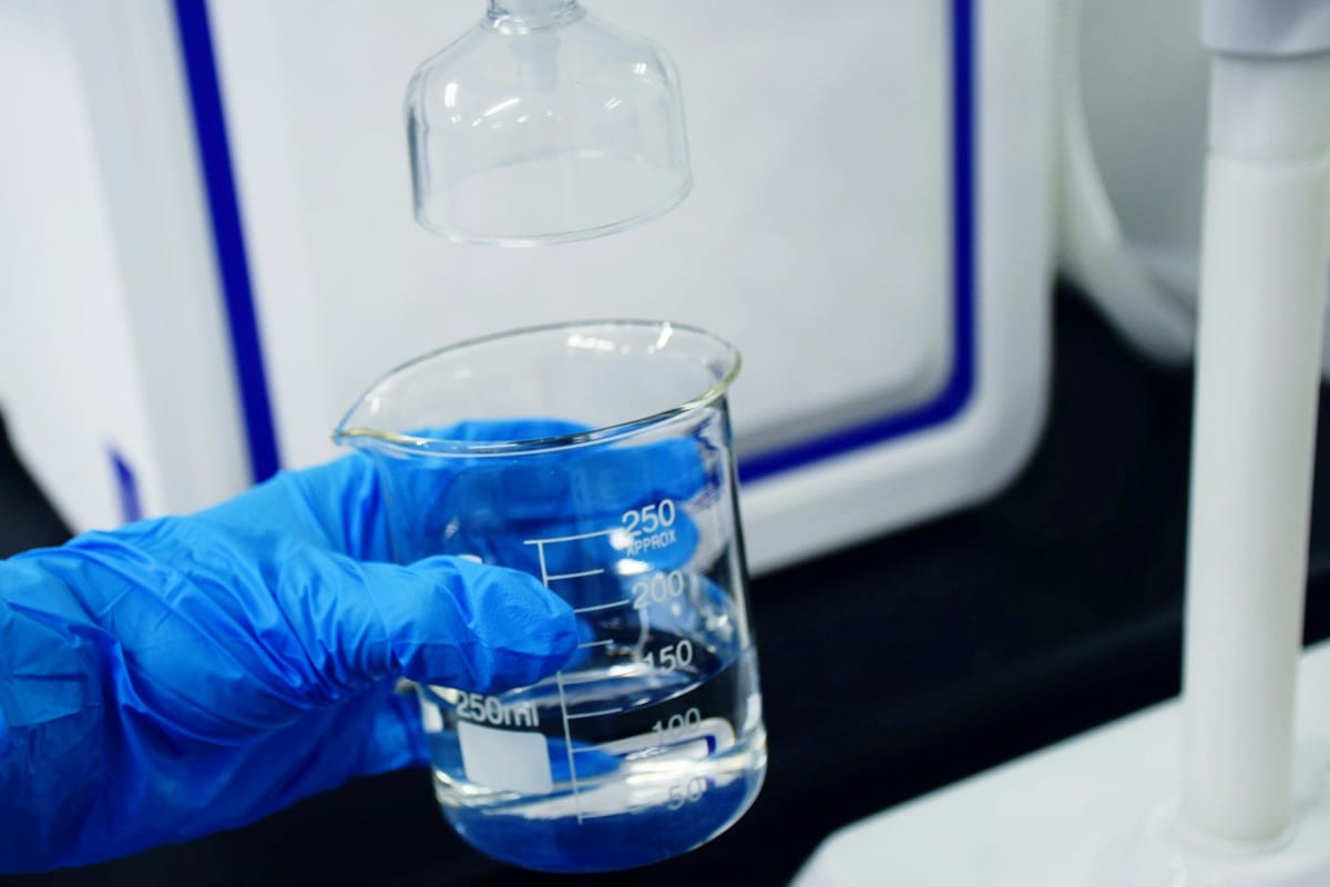 Blue-gloved hand holding a glass beaker of clear liquid beneath laboratory equipment.