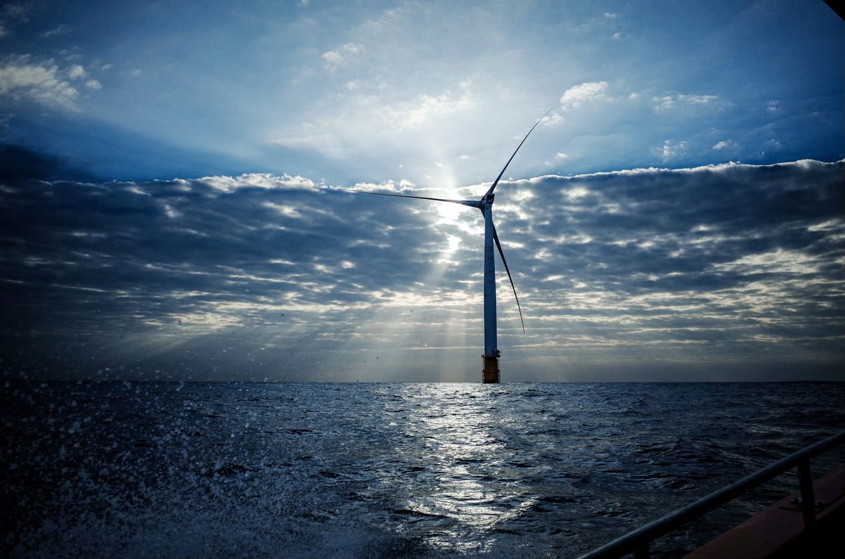 Offshore wind turbine rising from choppy ocean water, backlit by sunlight breaking through dramatic clouds.
