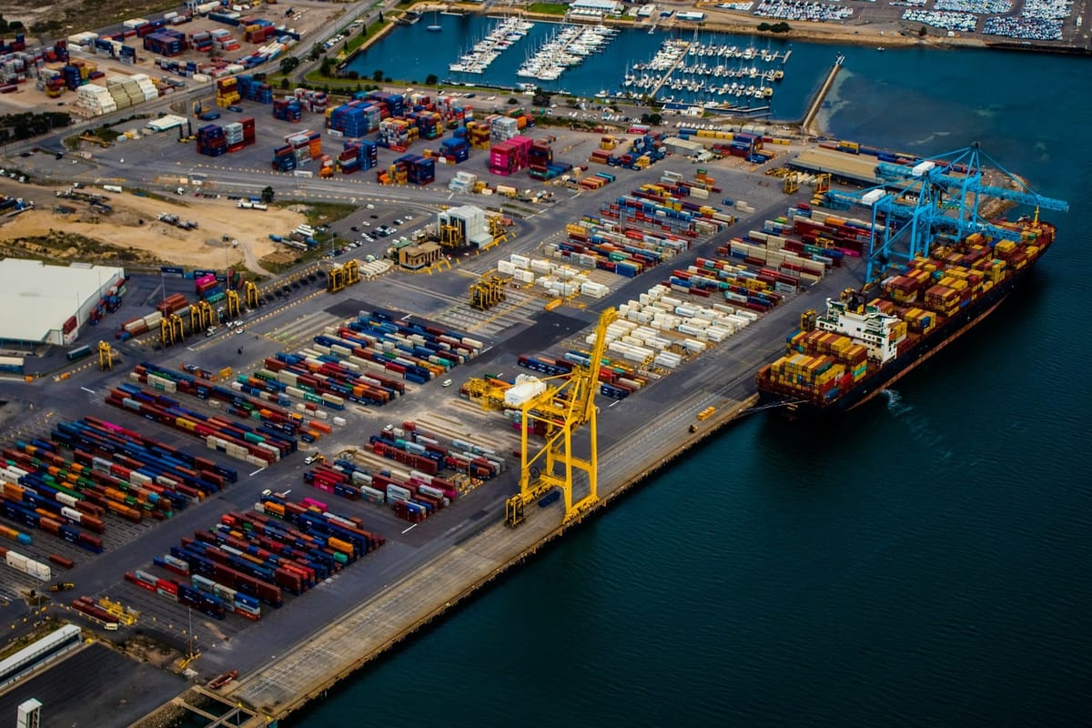 Aerial view of a container terminal with stacked shipping containers, cargo cranes, and a docked container ship beside a marina.