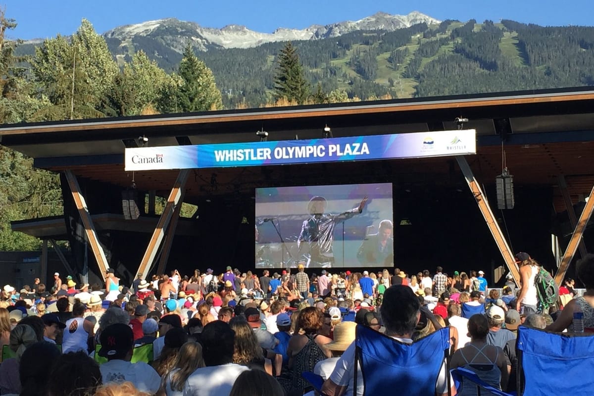 Crowd gathered at Whistler Olympic Plaza, with the stage and large video screen below Whistler Mountain on a clear summer day.