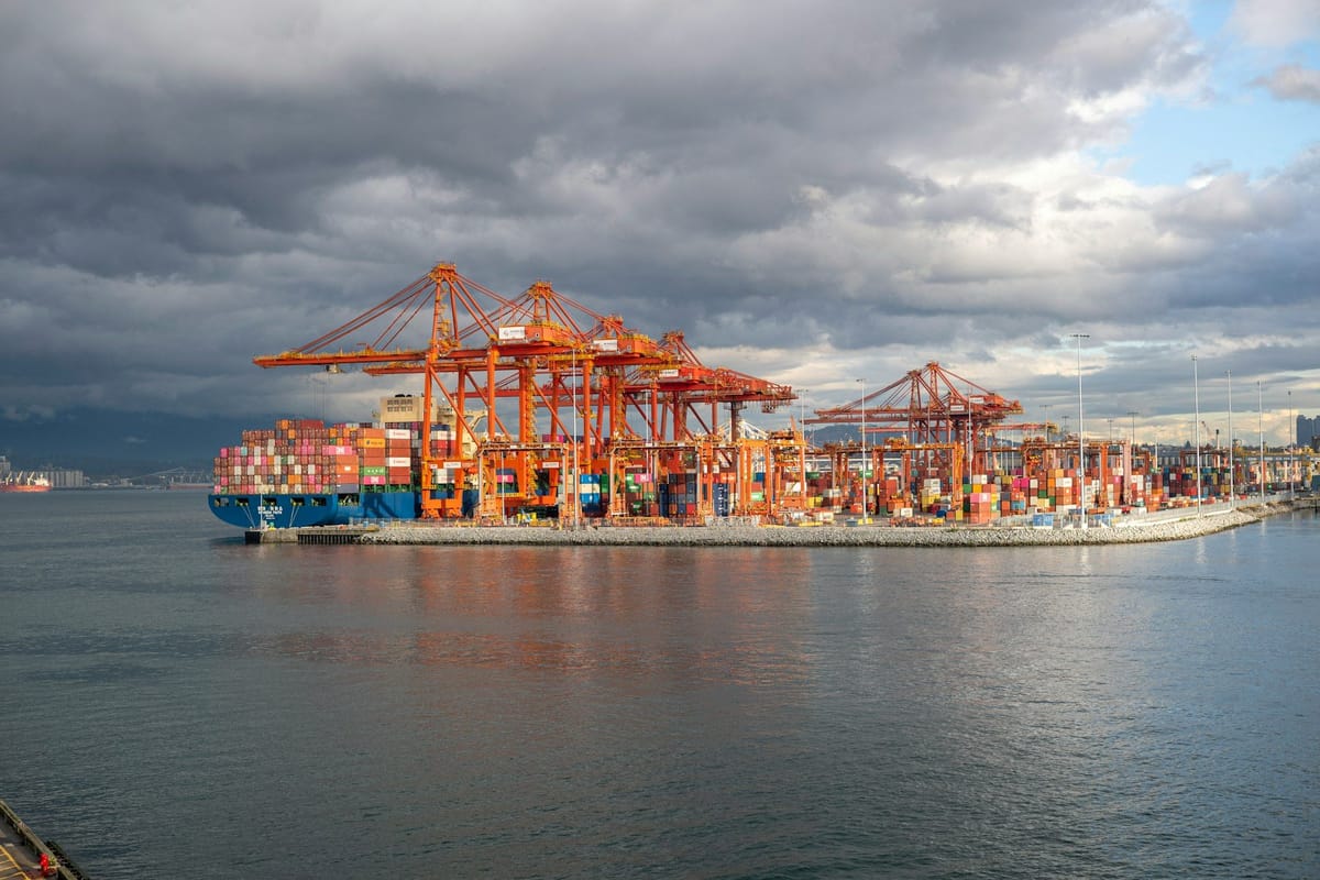 Container ship docked at a busy port terminal beneath large cargo cranes and dark storm clouds.
