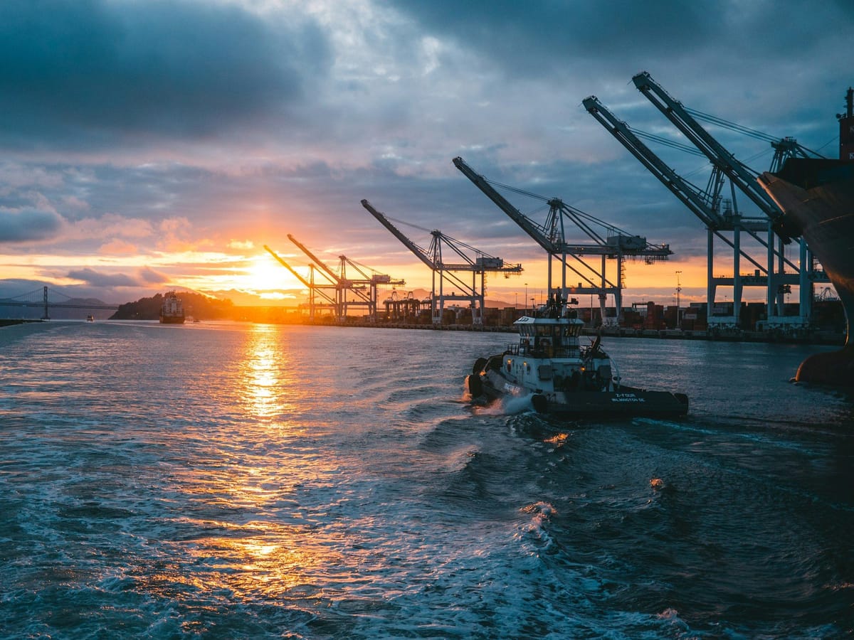 Tugboat moving through a busy container port at sunset, with cargo cranes lining the waterfront.