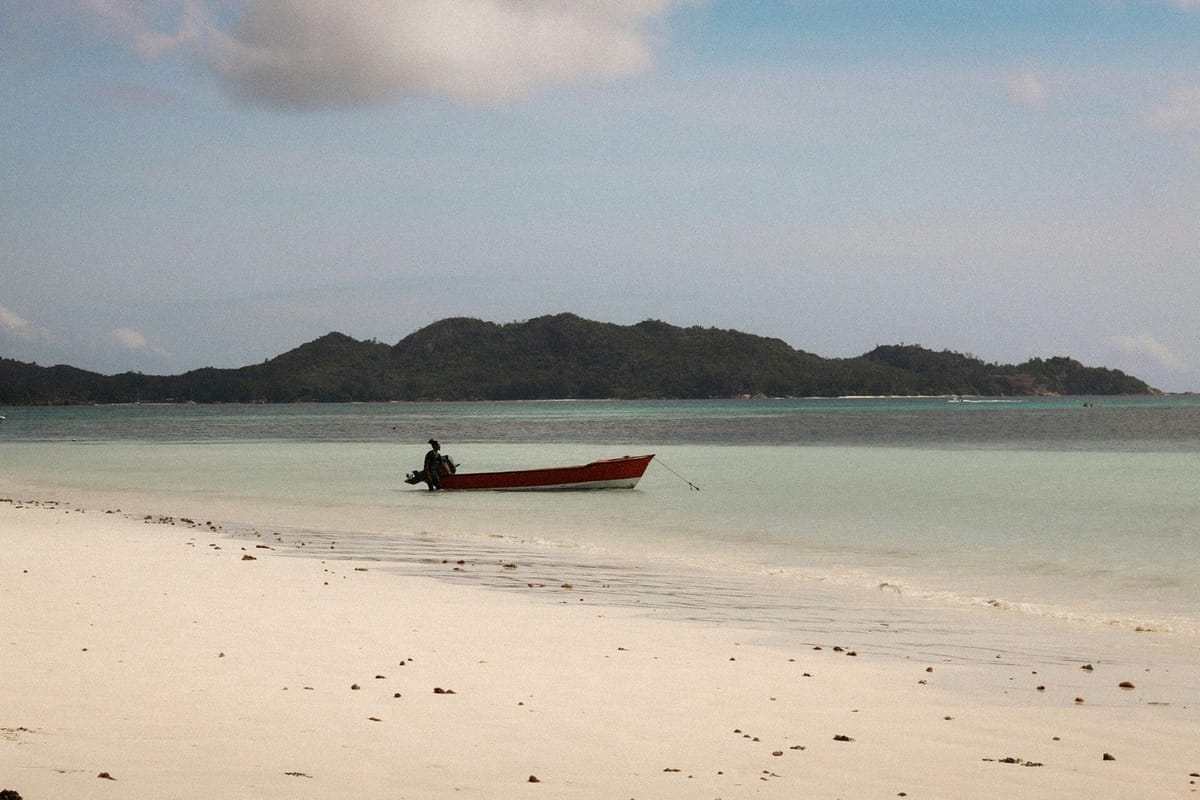 Small red motorboat anchored in shallow turquoise water beside a sandy beach, with a forested island in the distance.