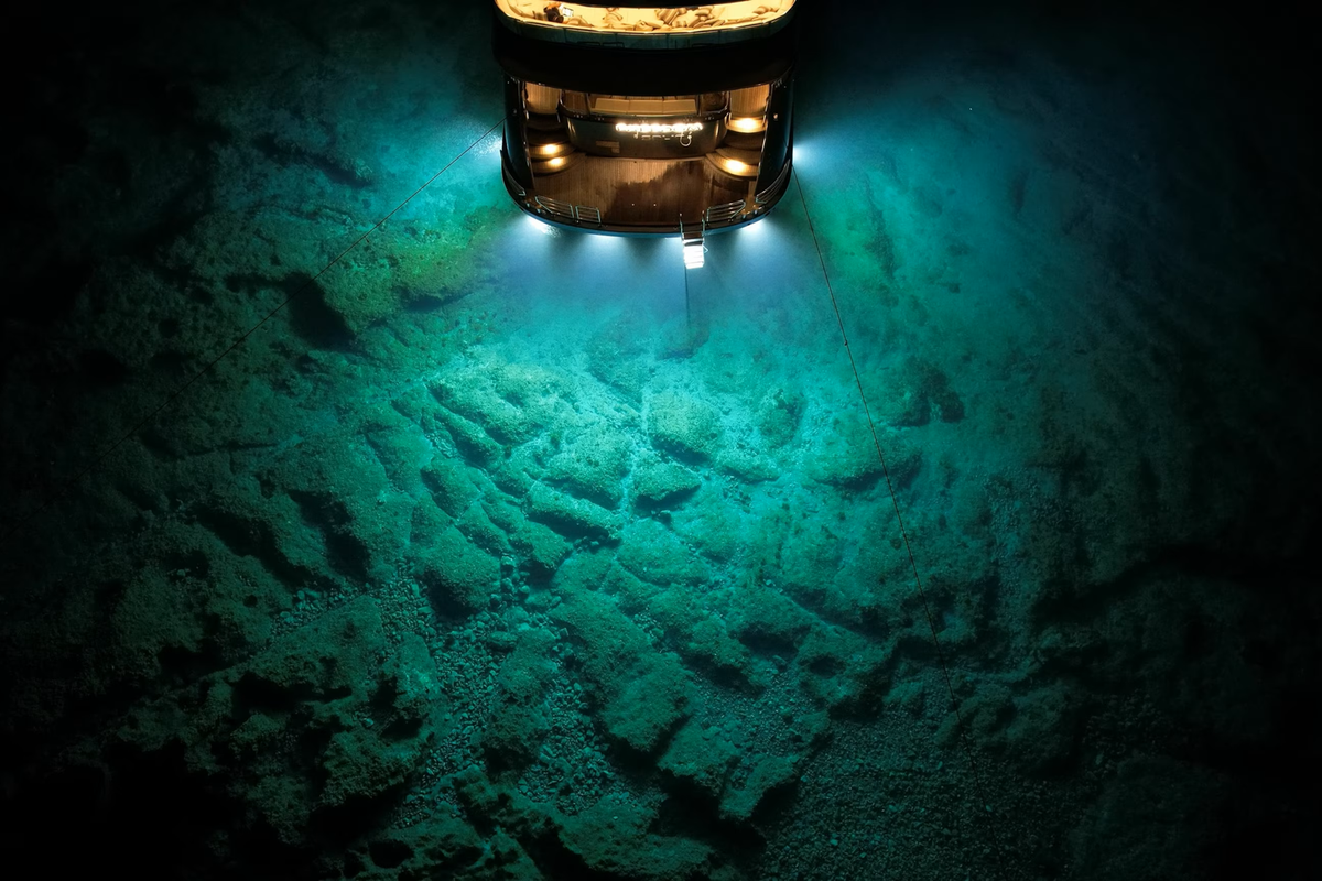 A research vessel at night illuminates the seafloor below with bright underwater lights, revealing rocky terrain in deep blue-green water.