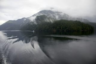 Mist-covered, forested mountains reflected in calm coastal waters as a small boat travels through the inlet.