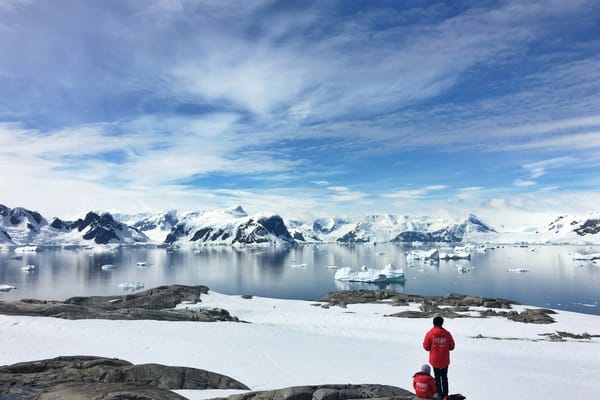 Two people in red parkas overlooking an Antarctic bay filled with icebergs and surrounded by snow-covered mountains.