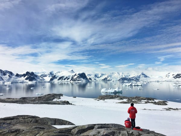 Two people in red parkas overlooking an Antarctic bay filled with icebergs and surrounded by snow-covered mountains.