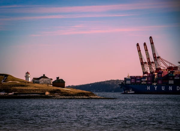 Container ship passing a lighthouse at the entrance to a harbour beneath a pink evening sky.