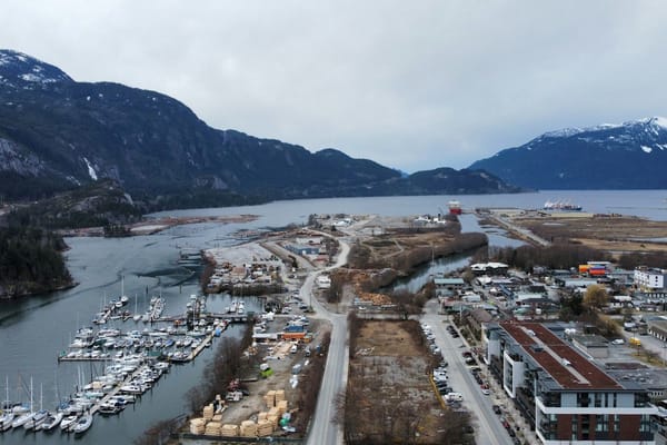 Aerial view of a marina and coastal industrial area surrounded by steep, snow-dusted mountains and a sheltered inlet.