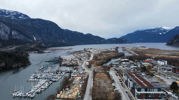 Aerial view of a marina and coastal industrial area surrounded by steep, snow-dusted mountains and a sheltered inlet.