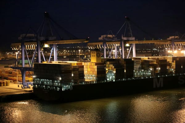 Container ship being loaded beneath illuminated cargo cranes at a port terminal at night.