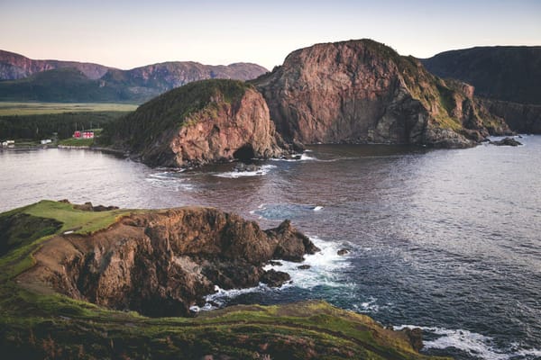 Rugged coastal cliffs surrounding a sheltered bay, with a small red house near the shoreline and mountains in the distance.