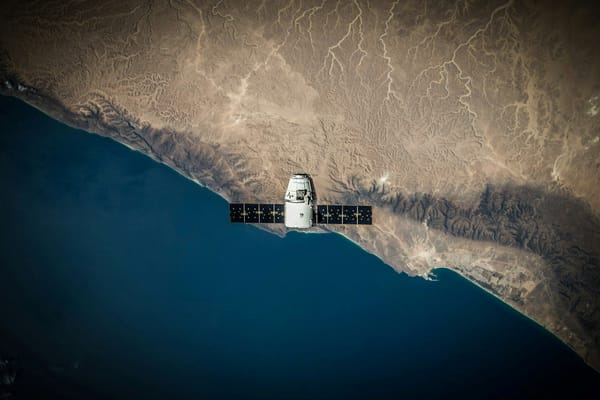 Spacecraft with solar panels orbiting above a rugged desert coastline and deep blue ocean.