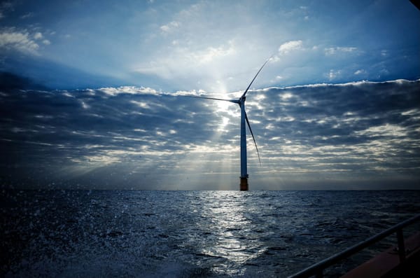 Offshore wind turbine rising from choppy ocean water, backlit by sunlight breaking through dramatic clouds.