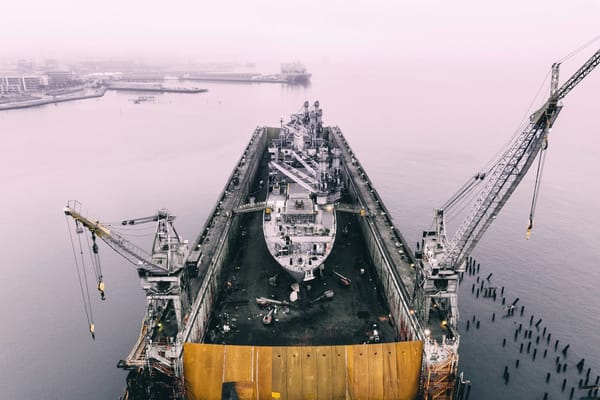 Aerial view of a large ship undergoing maintenance in a dry dock, surrounded by cranes in a foggy harbour.