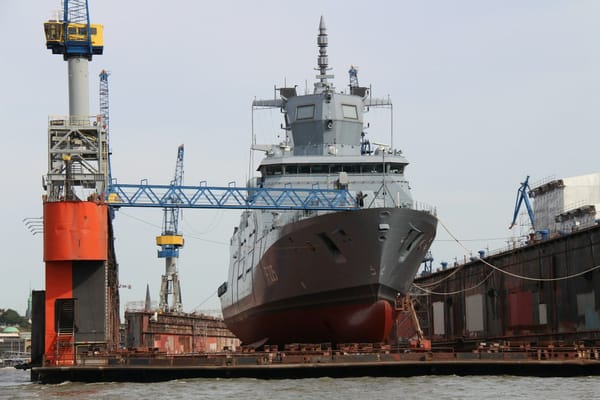 Large naval vessel undergoing maintenance in a dry dock, surrounded by cranes and shipyard equipment.