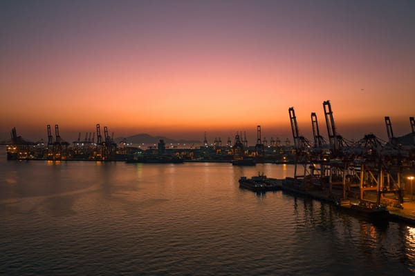 Container port illuminated at sunset, with cargo cranes silhouetted around a busy harbour.