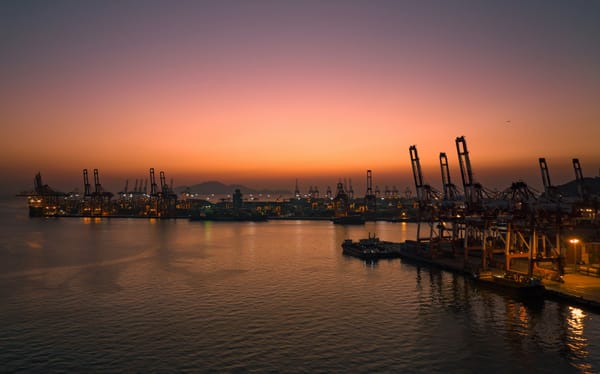 Container port illuminated at sunset, with cargo cranes silhouetted around a busy harbour.