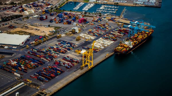 Aerial view of a container terminal with stacked shipping containers, cargo cranes, and a docked container ship beside a marina.