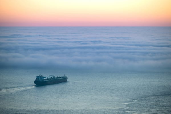 Cargo ship travelling beneath a low bank of fog under a softly coloured sunset sky.