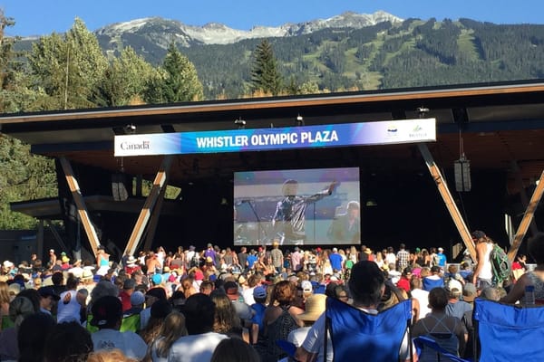 Crowd gathered at Whistler Olympic Plaza, with the stage and large video screen below Whistler Mountain on a clear summer day.