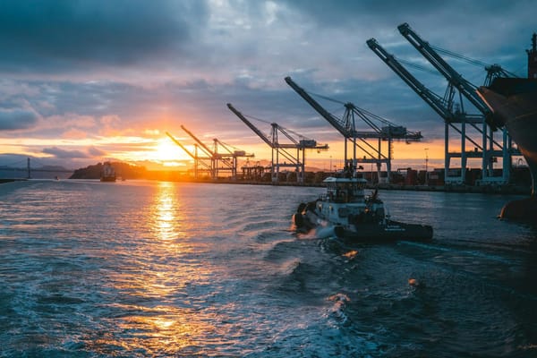 Tugboat moving through a busy container port at sunset, with cargo cranes lining the waterfront.