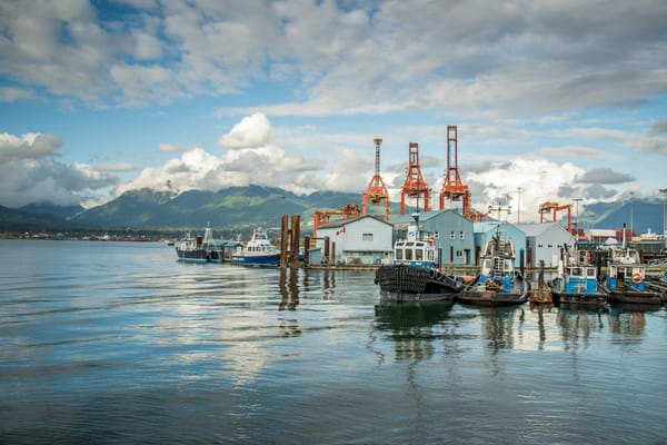 Tugboats moored at an industrial harbour, with container cranes and forested mountains in the background.