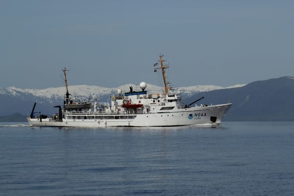 NOAA oceanographic research vessel underway in calm coastal waters, with snow-covered mountains in the background.