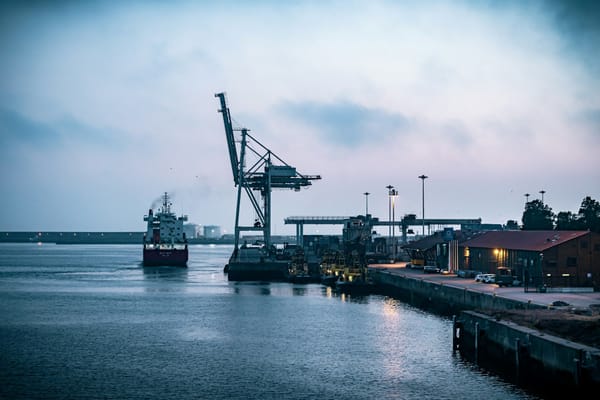 Cargo ship approaching a working port with cranes and waterfront buildings under a cloudy evening sky.