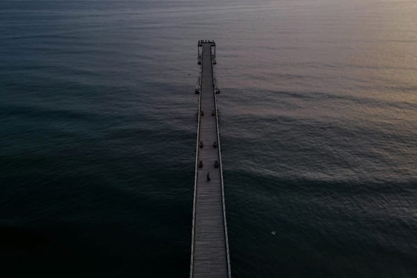Long wooden pier extending into calm ocean water at dusk, viewed from above.