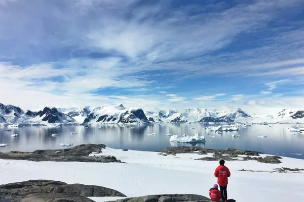 Two people in red parkas overlooking an Antarctic bay filled with icebergs and surrounded by snow-covered mountains.