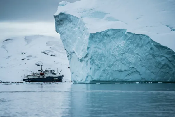Research vessel travelling beside a towering iceberg in icy polar waters.