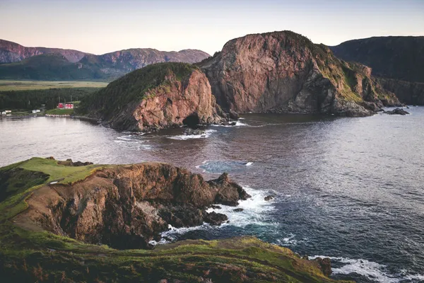 Rugged coastal cliffs surrounding a sheltered bay, with a small red house near the shoreline and mountains in the distance.