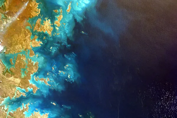 Satellite view of a rugged island coastline, with turquoise shallows fading into deep blue ocean.