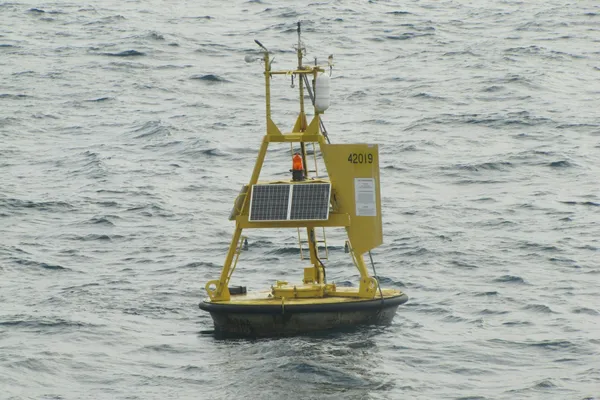 Yellow ocean-monitoring buoy with solar panels and instruments floating in choppy water.