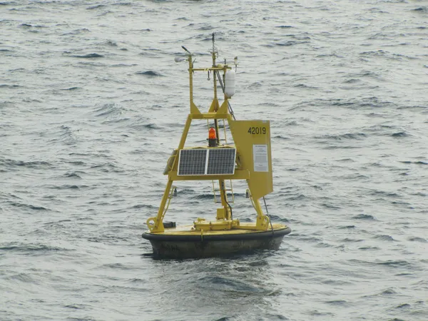 Yellow ocean-monitoring buoy with solar panels and instruments floating in choppy water.