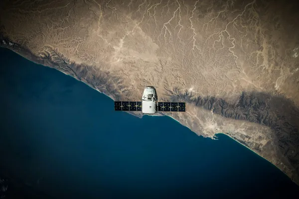Spacecraft with solar panels orbiting above a rugged desert coastline and deep blue ocean.