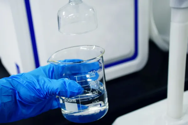 Blue-gloved hand holding a glass beaker of clear liquid beneath laboratory equipment.